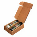 An open brown cardboard box containing two spice or herb seasoning bottles with black caps and colorful labels, photographed on a plain white background.